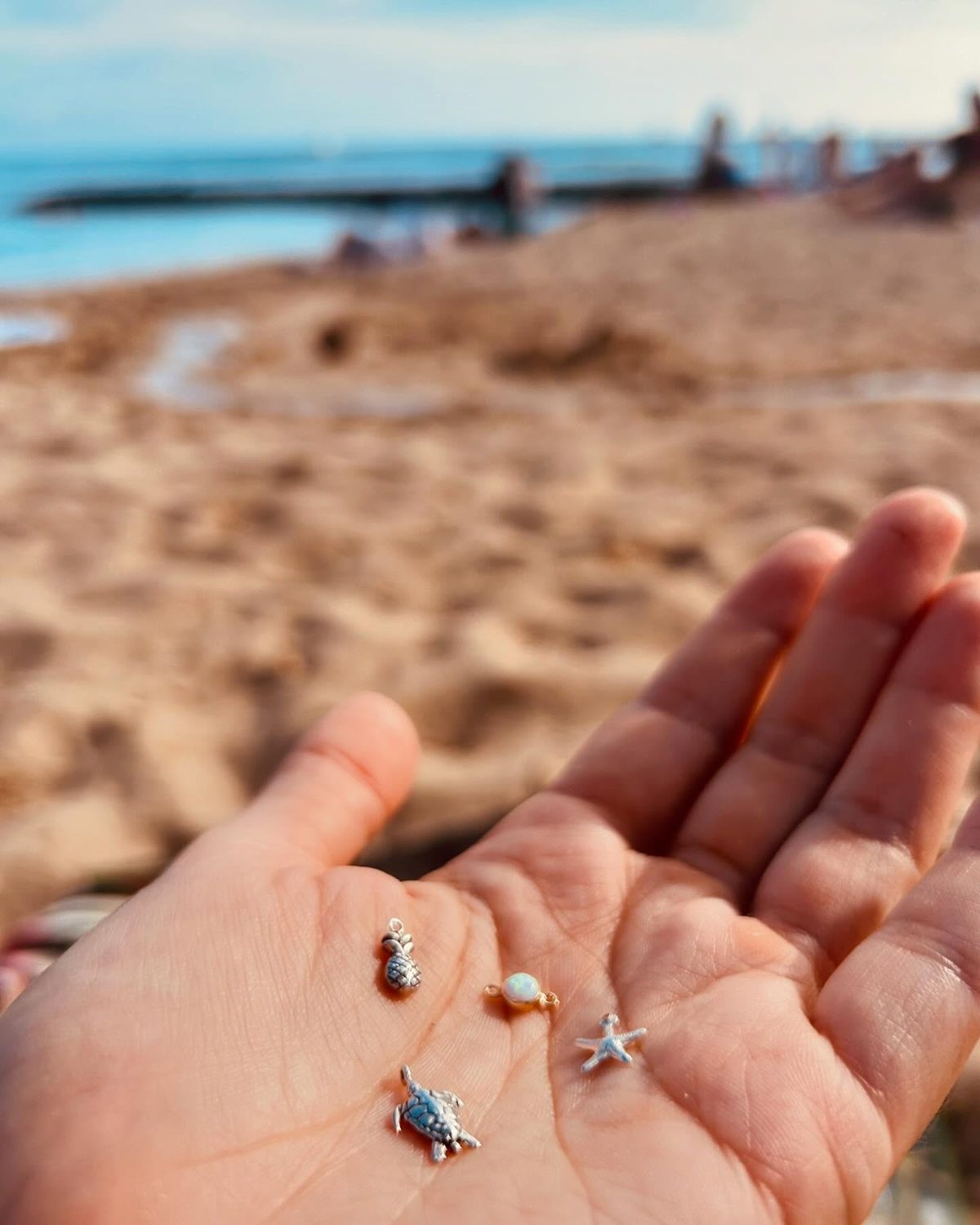 An open palm on the beach holding four tiny charms — a pineapple, an opal, a starfish and a sea turtle