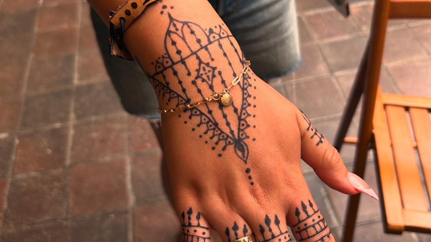 A gold chain bracelet with a tiny shell charm, worn on a hand decorated with henna